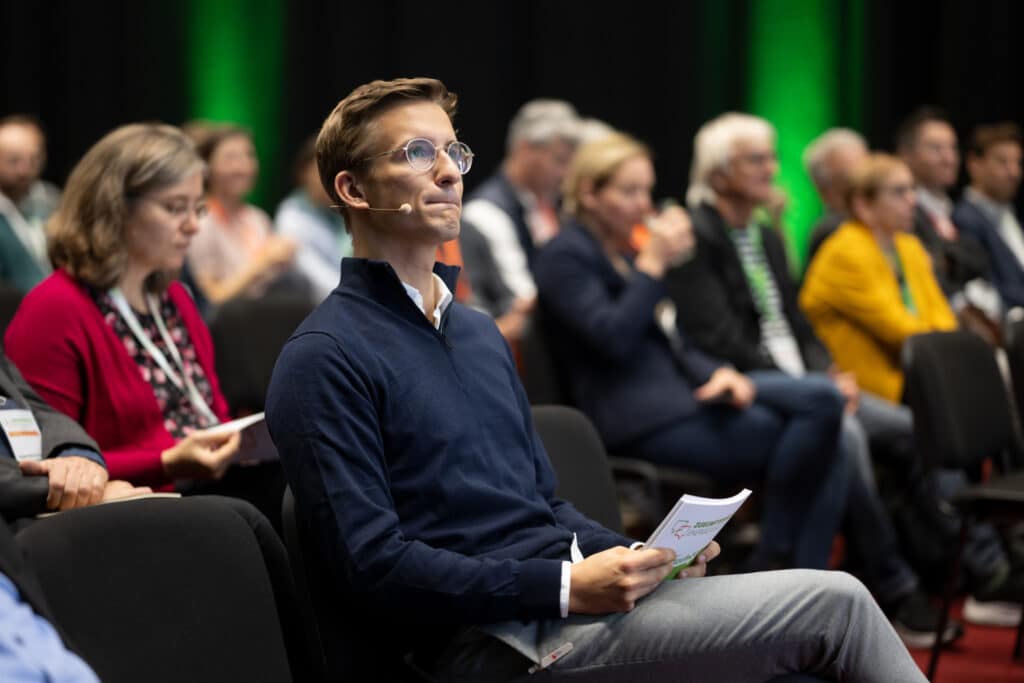 Moderator für Nachhaltigkeit sitzt im Publikum und beobachtet Panel auf der Bühne