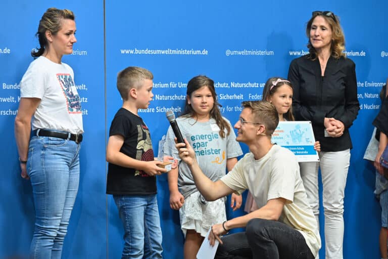 Moderator Julius Neumann kniet vor einem Kind, das er als Gewinner des Energiesparmeister-Wettbewerbs interviewt.
